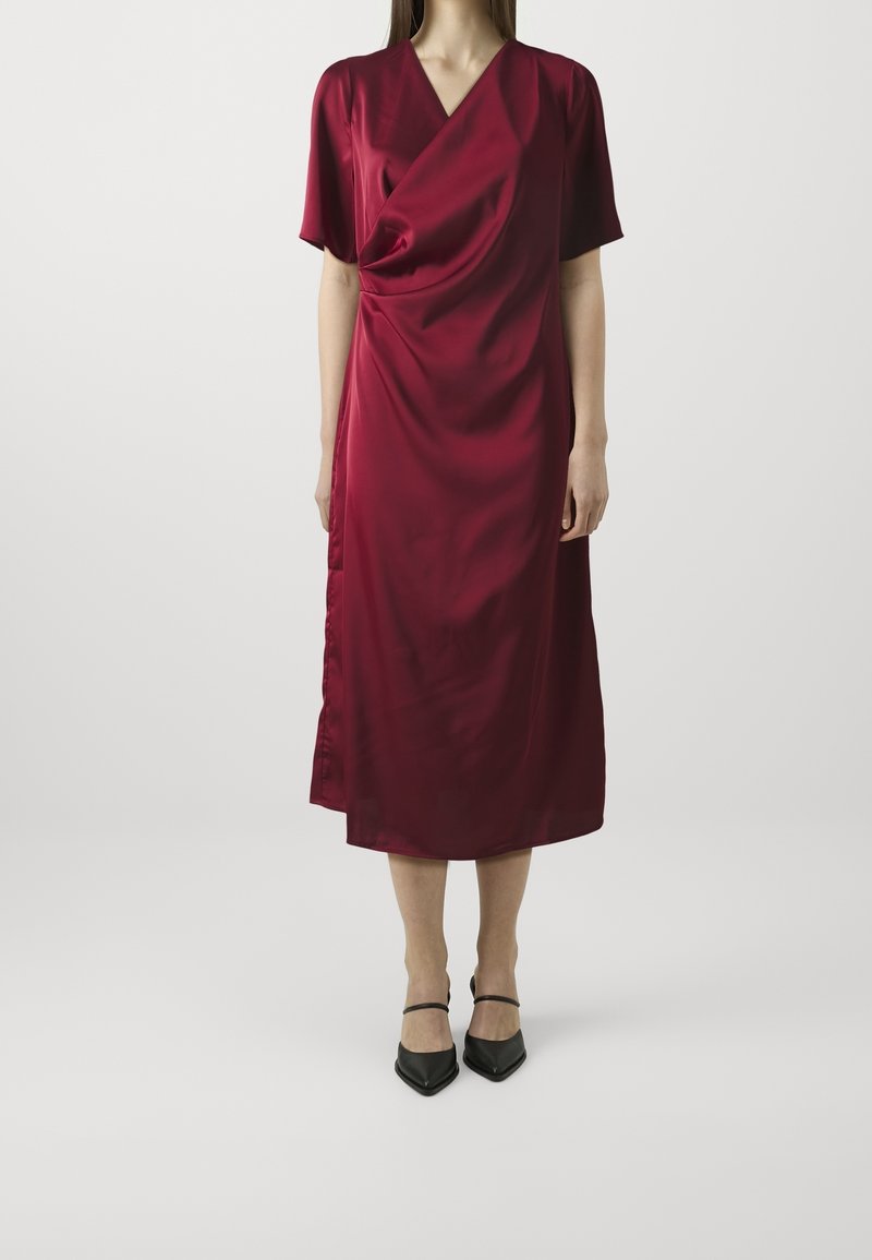 Femme portant une robe en satin rouge profond, à manches courtes et longueur genou, avec un devant drapé, et des escarpins noirs à bouts pointus avec des brides à la cheville.