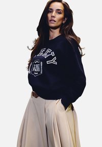 Sudadera azul marino con gráficos blancos, cuello redondo y mangas largas; combinada con una falda plisada beige clara y un cinturón marrón.