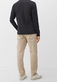 Camisa negra de manga larga con cuello redondo, combinada con pantalones beige claros y zapatillas blancas, mostrando la vista trasera. Los pantalones tienen bolsillos traseros.