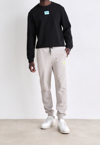 Sudadera negra con un logo azul, combinada con pantalones de chándal beige claro con un parche de logo amarillo y zapatillas blancas. Tela suave y corte relajado.