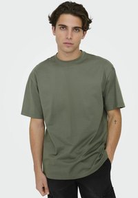 Olive green, short-sleeve t-shirt made of cotton, featuring a crew neckline and relaxed fit. Worn casually over black pants.