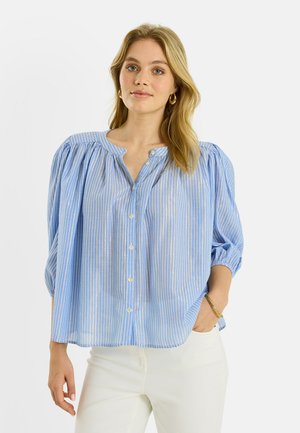 Femme aux cheveux blonds portant un chemisier rayé bleu et blanc à boutons et un pantalon blanc, debout avec une main dans la poche.