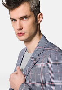 Szary marynarka w kratę z czerwonymi i niebieskimi liniami, z pojedynczym zapięciem na guzik i klapami typu notch. Noszona na szarym t-shircie z subtelnym akcentem w postaci logo.