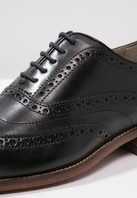 Derby en cuir noir avec des motifs perforés, lacets fermés, et une semelle en bois brun sur une surface blanche.