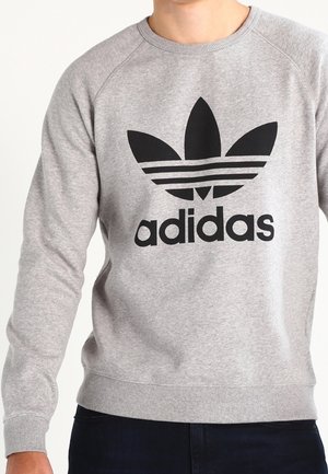 Grauer Sweater mit rundem Halsausschnitt, gerippten Bündchen und Saum, mit einem auffälligen schwarzen Adidas-Logo und drei schwarzen Streifen auf einem Blattdesign.