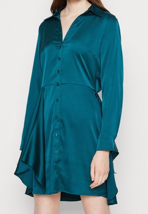 Sukienka z zielonym odcieniem morskim wykonana z atłasu, zapinana na guziki, z długimi rękawami, kołnierzem oraz asymetryczną falbaną po boku u dołu, noszona przez stojącą osobę.