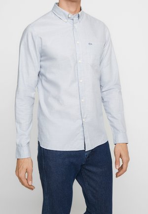 Homme portant une chemise boutonnée à manches longues bleu clair et un jean bleu foncé sur un fond uni.