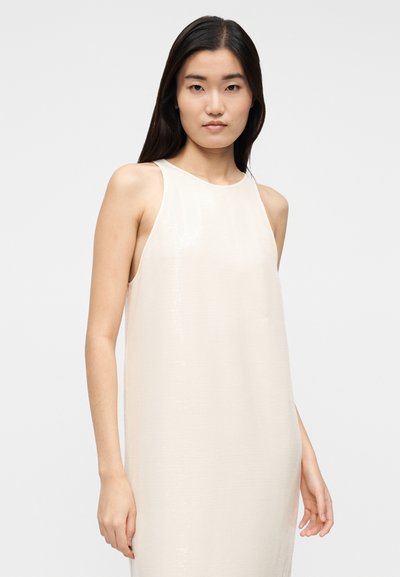 Ένα αμάνικο, ανοιχτό μπεζ φόρεμα από υφή ύφασμα, με άνετη γραμμή και διακριτική λάμψη, με σχεδιασμό πλαϊνών ανοίγμάτων στο τελείωμα.