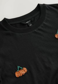 T-shirt en coton noir avec un col côtelé, présentant deux motifs de citrouilles en perles dans des nuances d'orange et de vert à l'avant.