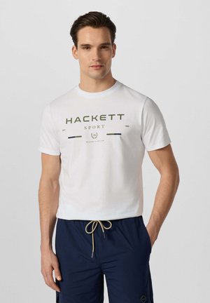 Hombre con camiseta blanca Hackett Sport y pantalones cortos azul marino con cordón, de pie contra un fondo liso.