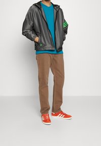 Man wearing gray zip-up jacket with green stripes on sleeve, teal shirt, brown pants, and orange Adidas sneakers standing with hands in pockets.