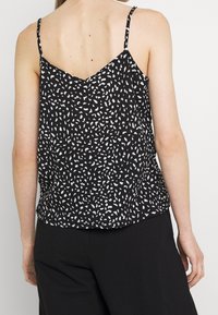 Top nero senza maniche con motivo astratto bianco, caratterizzato da spalline sottili e una schiena a forma di V. Il materiale sembra leggero e leggermente texture.