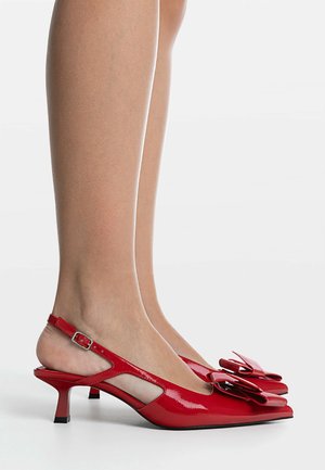 Red patent leather slingback heels with pointed toes and oversized decorative bows worn on feet against a plain background.