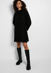 Schwarzes Kleid mit langen Ärmeln, gewelltem Saum und Laser-Cut-Details. Kombiniert mit kniehohen schwarzen Stiefeln. Strukturierter Stoff.