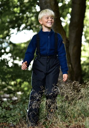 Blaue Fleecejacke mit Reißverschluss und schwarzen wasserdichten Overalls mit weißen Seitenstreifen. Die Person geht auf dem Gras zwischen Bäumen.