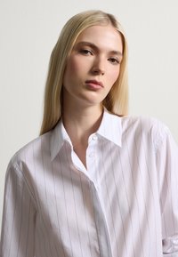 Camicia bianca con strisce verticali blu navy, dotata di colletto classico e tessuto morbido, con i bottoni parzialmente chiusi al collo.