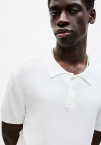 Hvid kortærmet polo t-shirt med en blød tekstur, der har en klassisk krave og en tre-knaps lukning. Diskrete stikninger er synlige.