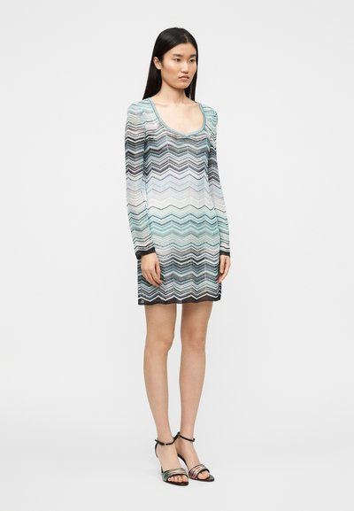 Φόρεμα με μακριά μανίκια και ζιγκ-ζαγκ μοτίβο σε αποχρώσεις του τιρκουάζ, λευκού και μαύρου. Κατασκευασμένο από ελαφρύ, διαφανές ύφασμα με στρογγυλή λαιμόκοψη.