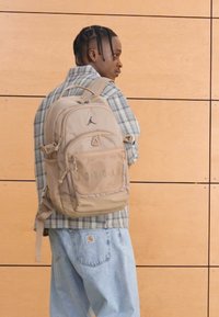 Mochila castanha com vários compartimentos, apresentando um logótipo preto na frente, usada por uma pessoa com camisa às riscas e jeans azul claro.