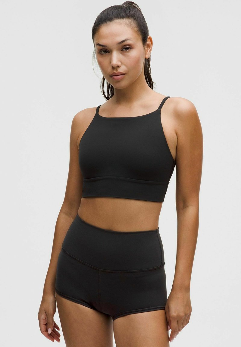 Frau trägt einen schwarzen Sport-BH und hoch taillierte Sportshorts, steht vor einem schlichten, hellen Hintergrund.