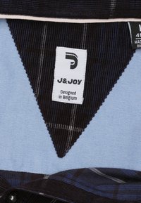 Donkerblauwe corduroy stof heeft een driehoekpaneel met een lichtblauwe achtergrond. Het label luidt "J&JOY" en "Ontworpen in België."