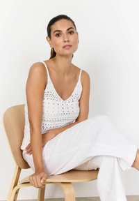 Bílý pletený top s výstřihem do V a otevřeným vzorem, v kombinaci s volnými bílými kalhotami, sedící na dřevěné židli u bílé zdi.