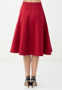 Jupe A-line rouge avec une taille ajustée, hem tombant sous le genou et texture lisse. Portée avec des chaussures à talons hauts noires.