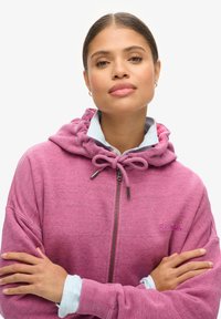 Haut à zip violet en tissu texturé, avec capuche à cordon et détail de logo sur la poitrine. Superposé sur une chemise rayée bleue.