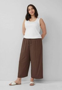 Weißes Tanktop, braune weit geschnittene Hose mit Kordelzugbund, strukturierter Stoff. Sandalen mit weißem Riemen und metallic Akzent.