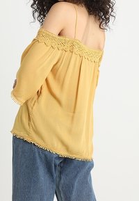 Yellow off-the-shoulder blouse with lace trim along the neckline and hem, featuring long sleeves and a loose, textured fabric design.
