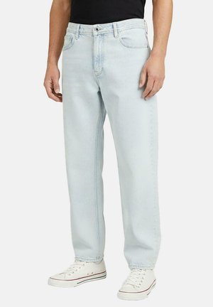 Mann trägt hellblaue Jeans mit geradem Bein, kombiniert mit weißen Sneakern und einem schwarzen Hemd, steht vor einem schlichten Hintergrund.