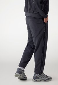 Person, der schwarze Jogginghosen mit Streifen "GUESS ATHL DEPT" und grauen Sneakers trägt, steht mit einer Hand in der Tasche gegen einen weißen Hintergrund.