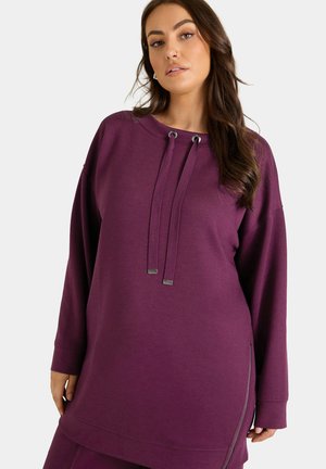 Kvinde iført en lilla langærmet pullover med snore og sidesyning, stående mod en ensartet lys baggrund.