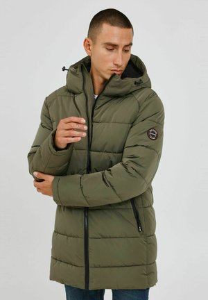 Chaqueta acolchada verde oliva con capucha, cierre de cremallera en la parte frontal y bolsillos laterales. Presenta un parche redondo con el logo en la manga izquierda. Textura suave.