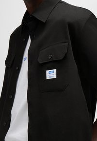 Zwart overhemd met knopen en twee borstzakken, voorzien van een wit label met blauwe tekst. Gedragen over een wit T-shirt. Gladde stoftextuur.