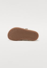 Suela de goma marrón con surcos texturizados, que presenta patrones circulares. El exterior tiene un acabado beige con un detalle de correa ajustable.