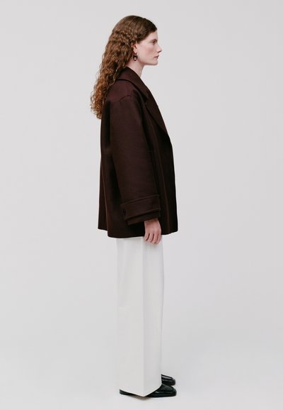 Cappotto oversize marrone con colletto a sottopunto, indossato sopra pantaloni bianchi a gamba larga e scarpe nere. I capelli sono ricci. Vista di profilo su uno sfondo neutro.