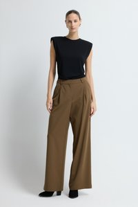 Schwarzes, figurbetontes Oberteil mit Schulterpolstern, kombiniert mit hoch taillierter, weit geschnittener brauner Hose aus glattem Stoff. Schwarze Stiefel mit Absatz.