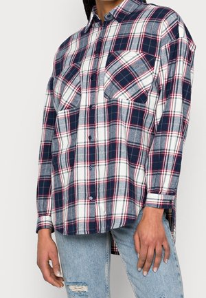 Person trägt ein locker sitzendes kariertes Hemd in Marineblau, Weiß und Rot mit Brusttaschen, kombiniert mit hellblauen, zerrissenen Jeans.
