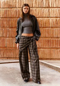 Mlajša ženska pozira ob teksturirani opečni steni, nosi sive rebraste kratke pulover s polo ovratnikom, črno usnjeno jakno in široke kariraste hlače z ovitkom.