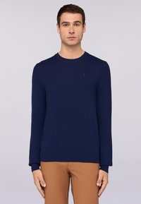 Granatowy sweter dziany z długimi rękawami, okrągłym dekoltem i ściągaczami przy rękawach. Ma małe logo na piersi. Noszony z beżowymi spodniami.