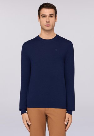 Suéter de punto azul marino con mangas largas, cuello redondo y puños acanalados. Presenta un pequeño logo en el pecho. Se lleva con pantalones beige.