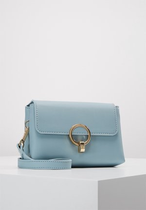 Lyseblå læderclutch med gyldent cirkulært ringlukning og aftagelig håndledsrem, placeret på en hvid fod på en ensfarvet baggrund.