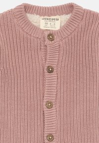 Svetlo roza pletena jopica z okroglo ovratnico, ki ima mehak notranji podlog in pet lesenih gumbov spredaj. Oznaka velikosti vidna.