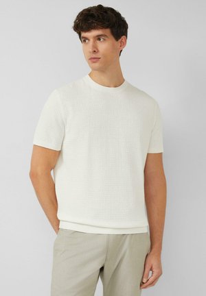 Młody mężczyzna w białym swetrze z krótkim rękawem o fakturowanej powierzchni oraz jasnobeżowych spodniach, stoi z rękami za plecami, patrząc w lewo.