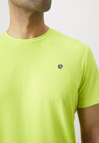 Mees, kellel on seljas eredat laimi roheline spordisärk, millel on väike must logo vasakul rinnal, lähedane vaade ülemisele kerele ja kaelale.