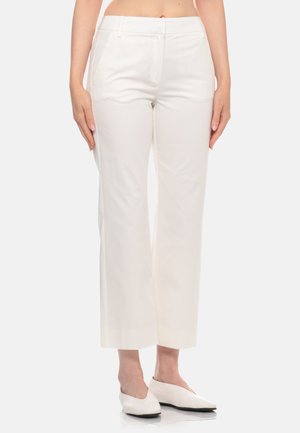 Donna che indossa pantaloni bianchi a vita alta e taglio dritto e scarpe bianche basse, in piedi con le braccia rilassate ai lati.