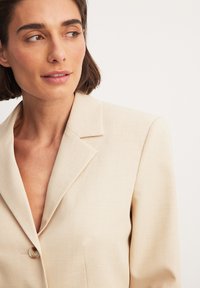 Beige blazer met een insnoering, sluiting met één knoop en een gestructureerde stof. Beschikt over een borstzak en een getailleerde pasvorm.