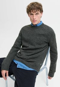 Pull en maille gris foncé avec un col rond, porté sur une chemise à col bleu clair. Le tissu a une apparence texturée et des poignets côtelés.