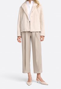 Veste en fausse fourrure beige avec col châle, associée à un pantalon beige sur mesure et des escarpins slingback à bout pointu, mettant en valeur un design épuré.